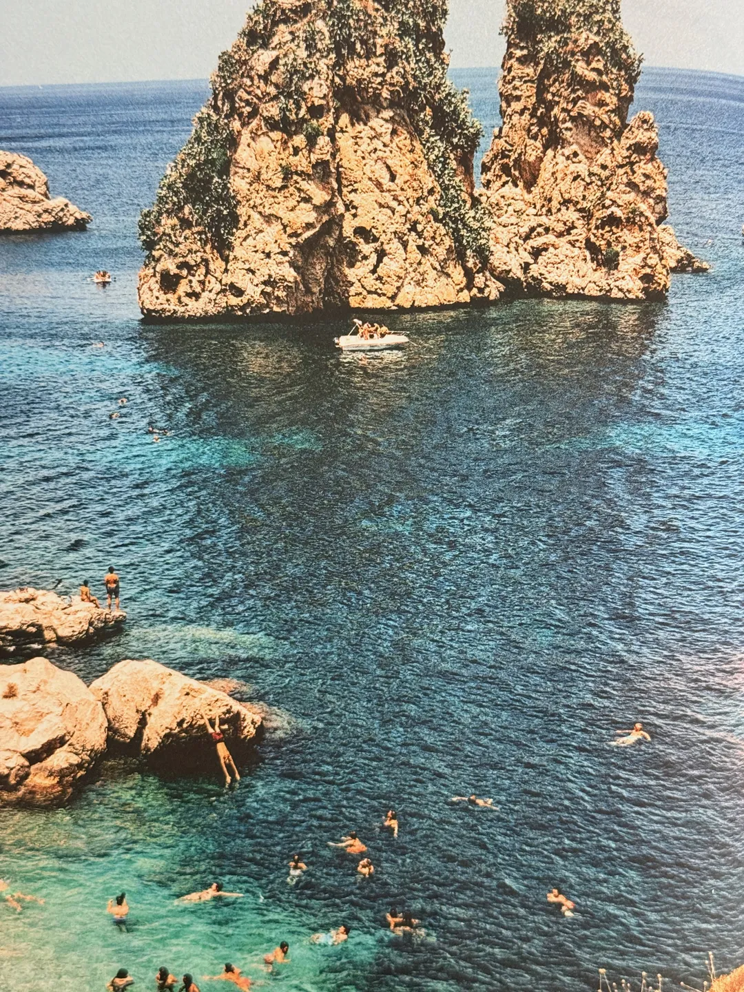 Swimmers in clear water around tall sea stacks off the Sicilian coast